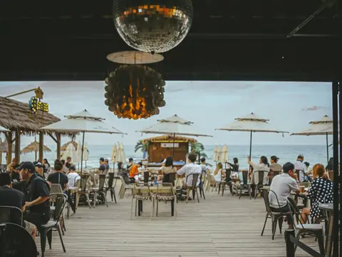 Mood photo (not the venue): Guests at tables on a wooden deck by the beach